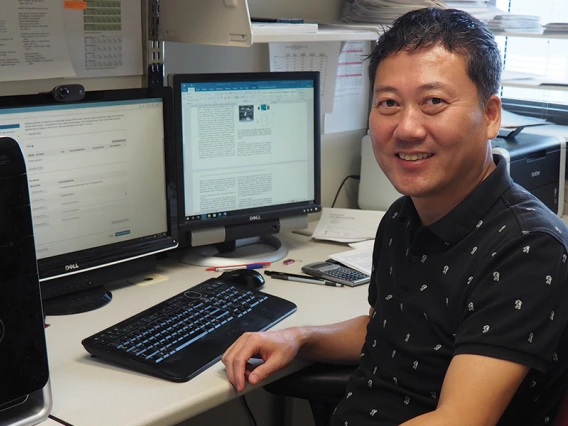 A man in a black short-sleeve shirt poses in front of two computer monitors.