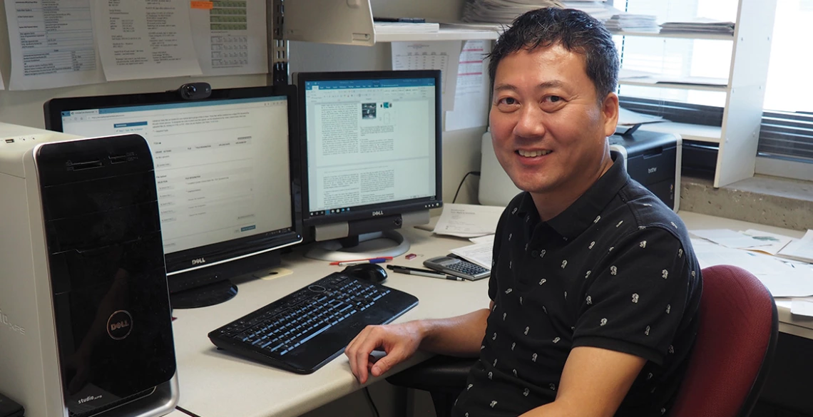 A man in a black short-sleeve shirt poses in front of two computer monitors.