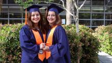 Two women in matching graduation caps and gowns hold hands in a courtyard.
