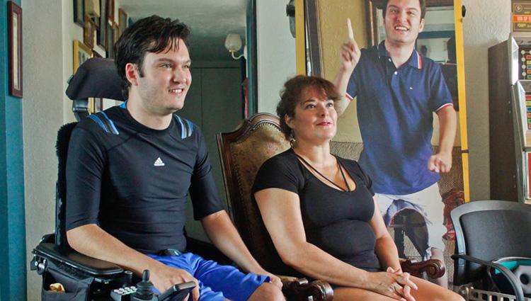 Jeffrey Bristol and his mother, Hermalinda, sit in their living room in Tucson. Jeffrey is wearing a dark blue athletic shirt and bright blue exercise shorts, sitting in a padded electric wheelchair. 