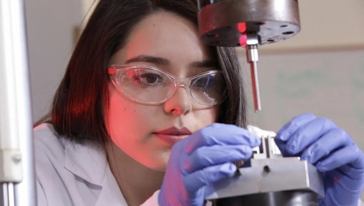 A woman wearing a lab coat and goggles focuses her attention on a small metal object.