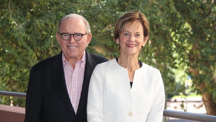 Peter and Nancy Salter stand together next to a railing in front of leafy green trees.