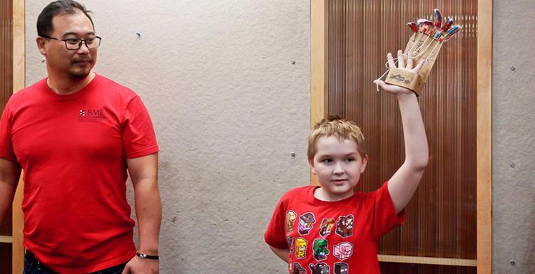A man standing next to a boy raising his hand, which is in the glove of a mechanical hand.