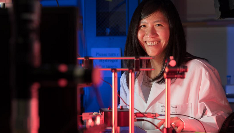 Judith Su wearing a lab coat and smiling. There is a sensor setup made of metal rods on the table in front of her.