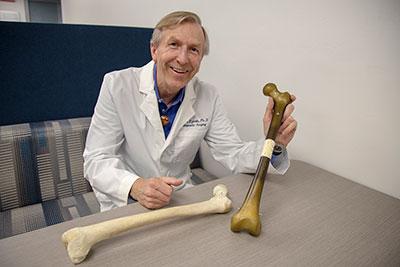 A man in a white lab coat posing with two large bones while sitting at a desk