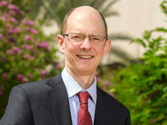 A bespectacled man in a suit and tie smiles for a portrait.
