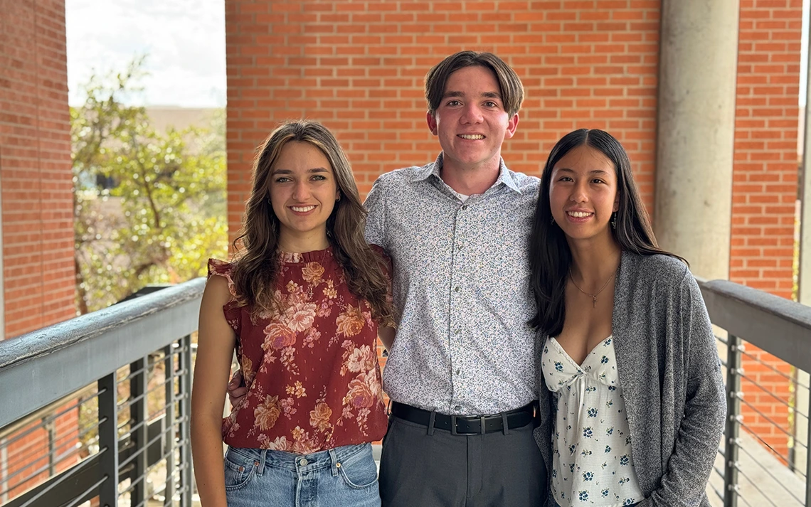 Three people stand in front of a brick wall.