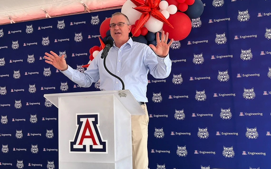 A man in a light blue shirt makes a speech at a white podium.