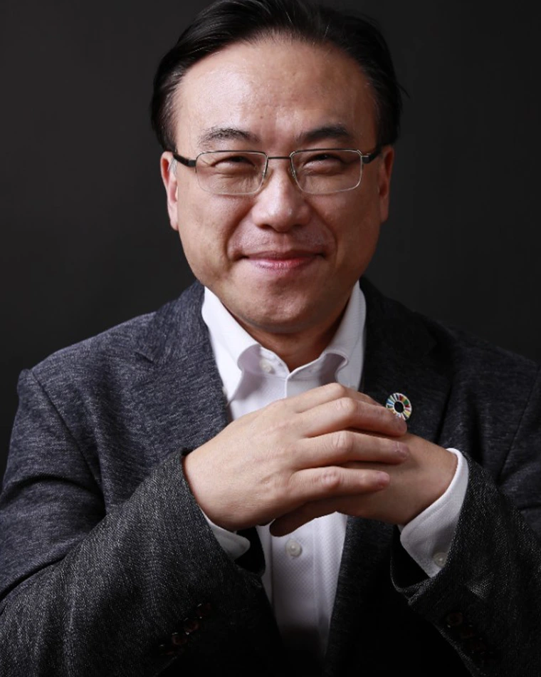 person with short, dark hair, wearing glasses, dark suit and white shirt with hands folded