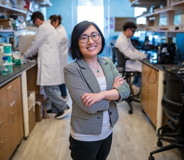 person with short dark hair and glasses, standing in lab, arms crossed
